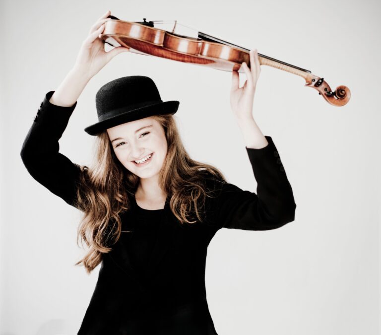 Een meisje in een zwarte jurk en hoed houdt een viool boven haar hoofd en glimlacht.