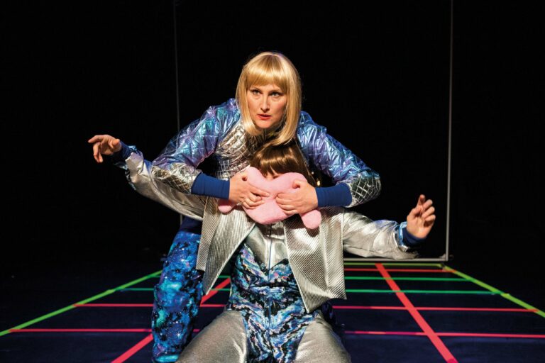 Twee performers in futuristische outfits poseren op een felgekleurde achtergrond, waarbij de ene achter de ander staat en een knuffel vasthoudt.