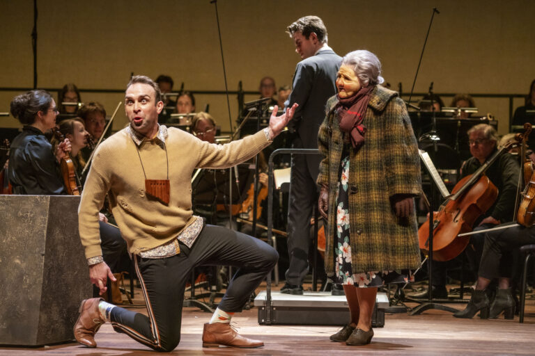 Een man knielt en praat met een oudere vrouw in een schouwburgsetting, terwijl een muzikant op de achtergrond aanwezig is.