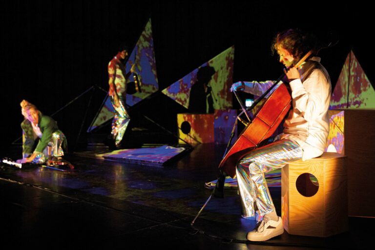 Twee muzikanten voeren op het podium, met kleurrijke geometrische vormen op de achtergrond en één muzikant die een cello bespeelt.