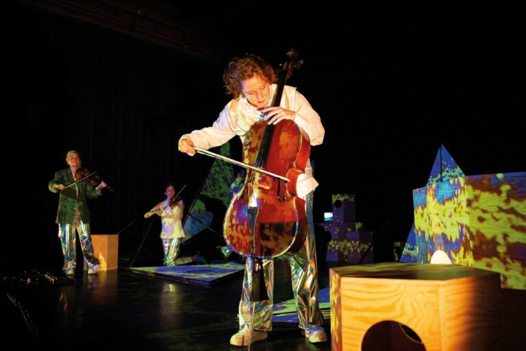 Een performer speelt cello op een podium, omringd door kleurrijke decorstukken.