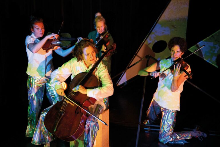 Een groep muzikanten speelt op een podium, waarbij een cellist centraal staat en andere leden violen en fluiten bespelen.