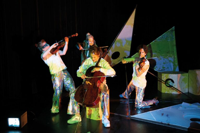 Een muzikale voorstelling met vier performers die instrumenten spelen op een kleurrijk podium.