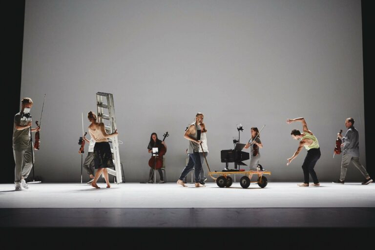 Een groep dansers en muzikanten op het podium, met een ladder en een wagen als decor.