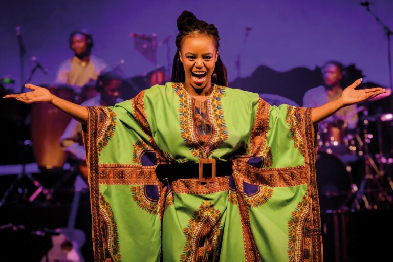 Een vrouwelijke artiest in een kleurrijke groene outfit zingt en uitdrukt veel enthousiasme op het podium.