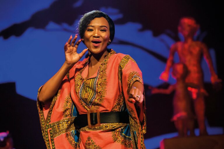 Een lachende performer in een kleurrijke outfit zingt op het podium met een dynamische achtergrond.