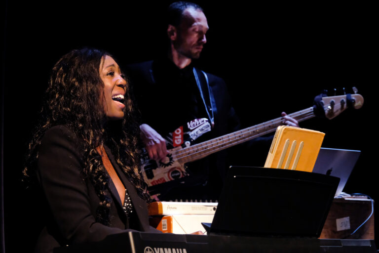 Een vrouw speelt piano en zingt terwijl een man met een basgitaar op de achtergrond staat.