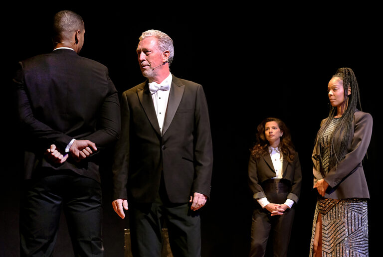 Vier acteurs in formele kleding staan op het podium tijdens een toneelstuk.