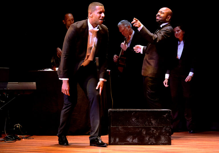 Een groep acteurs in formele kleding voert een theatrale scène op een podium uit.