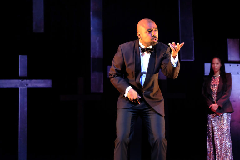 Een man in een zwart tuxedo voert een theatrale monoloog op een podium.