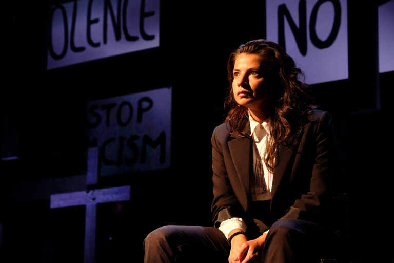 Een vrouw in een kostuum zit op een podium te midden van protestborden met boodschappen over geweld en racisme.