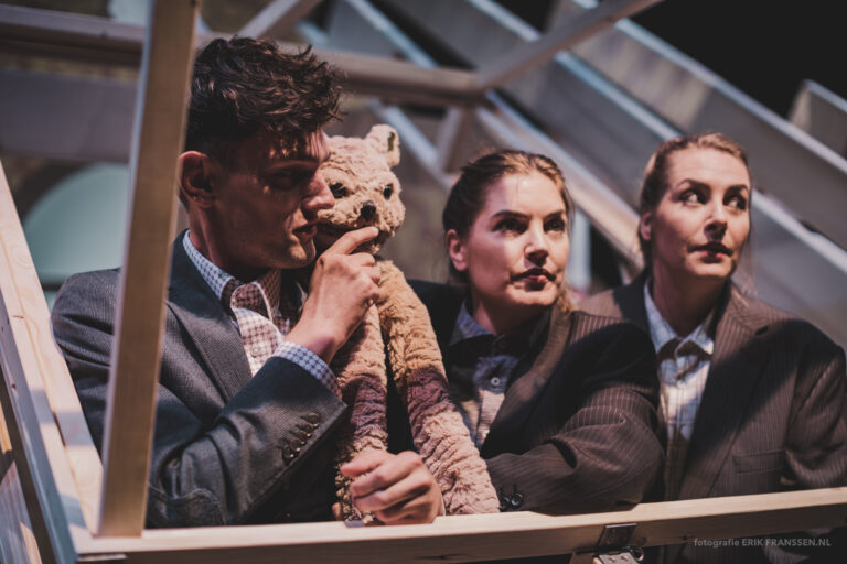 Drie acteurs in formele kleding kijken bezorgd terwijl één van hen een knuffelbeer vasthoudt in een theatrale setting.