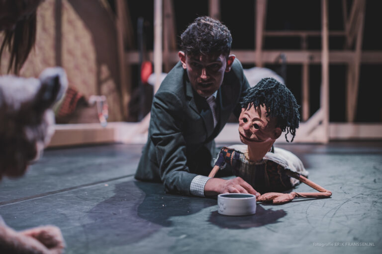 Een man in een pak ligt op een podium en interacteert met een uit hout gemaakte pop met een krullend kapsel.