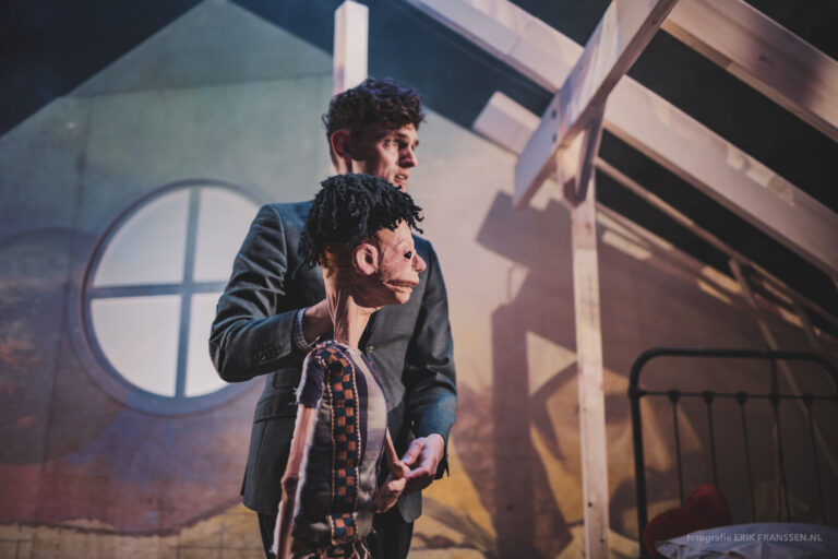 Een poppenspeler houdt een marionet vast op een podium met een decor op de achtergrond.