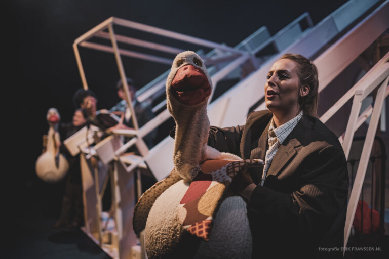 Een toneelacteur houdt een grote handpop van een gans vast terwijl andere acteurs op de achtergrond bezig zijn.