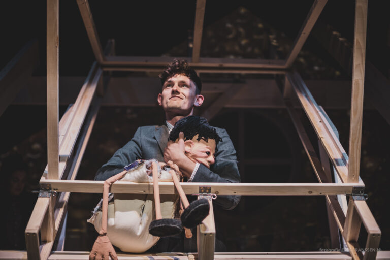 Een man in een grijs kostuum staat op een platform en houdt een pop vast.