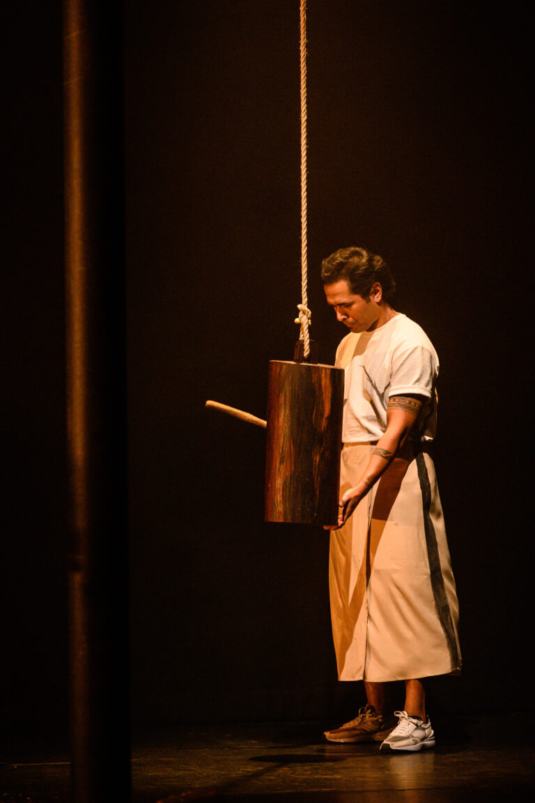 Een man in een witte outfit slaat met een stok op een hangende houten cilinder in een donkere omgeving.