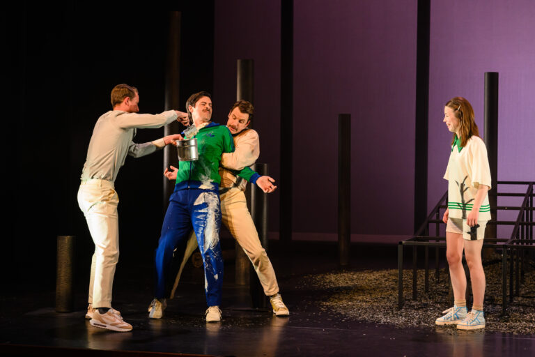 Drie acteurs zijn op het podium met elkaar in interactie, terwijl een vierde acteur op een stoel zit.