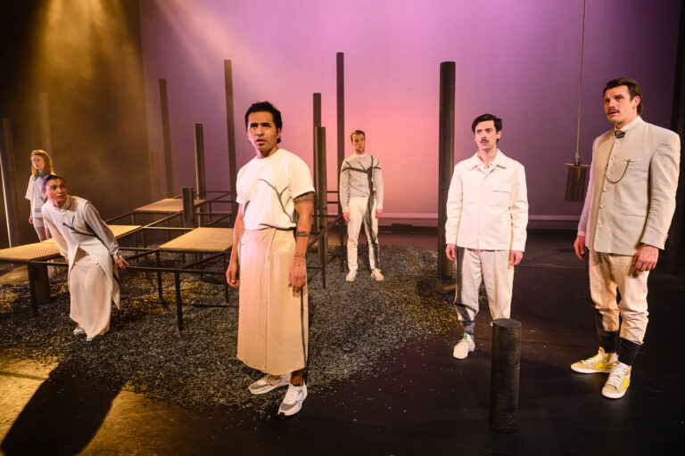 Een groep vier acteurs in witte kostuums staat op een podium omringd door houten palen en tafels onder kleurrijke verlichting.