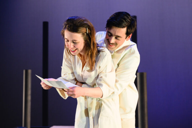 Twee acteurs in een theaterstuk, met de vrouw die een tekst leest terwijl de man haar van achteren omhelst.