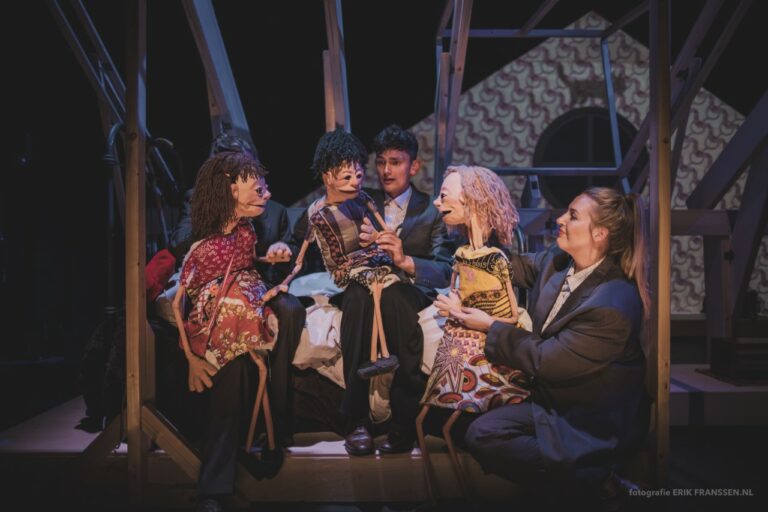 Een groep mensen in kostuums zit samen op een podium, omringd door decor en rekwisieten.