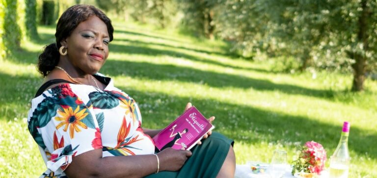 Surinaamse vrouw met zonnebril op haar hoofd, een boek in haar handen en kleurrijke jurk op een grasveld.
