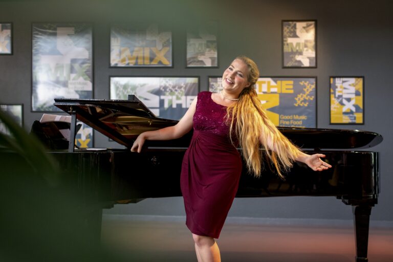 Een persoon poseert in een elegante jurk naast een piano in een artistieke setting.