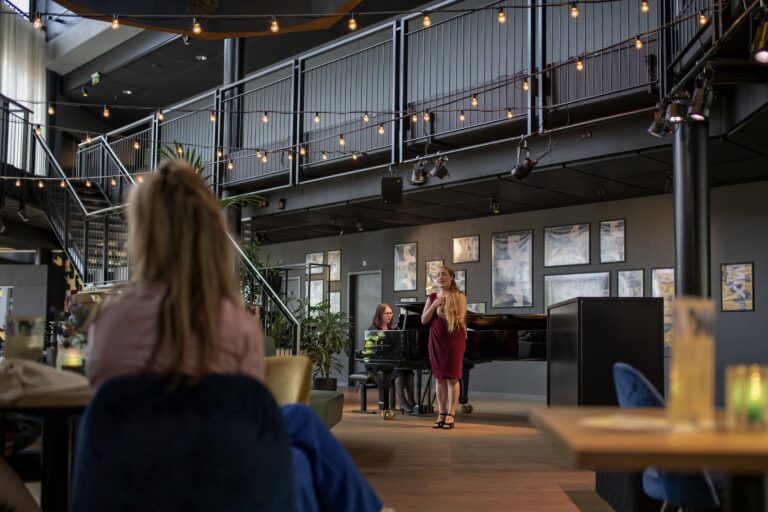 Een vrouw in een rode jurk zingt op een podium in een moderne ruimte, terwijl een klein publiek kijkt.