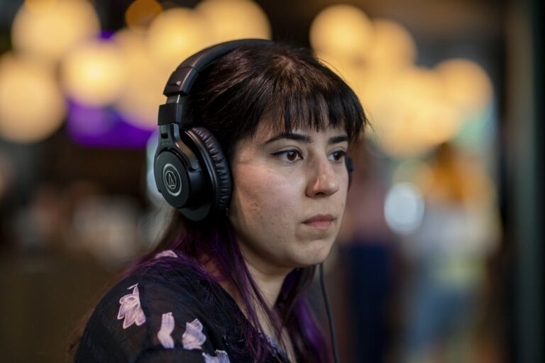 Een jonge vrouw met hoofdtelefoon kijkt geconcentreerd in de richting van de camera, met een vervaagde achtergrond van lichtbollen.