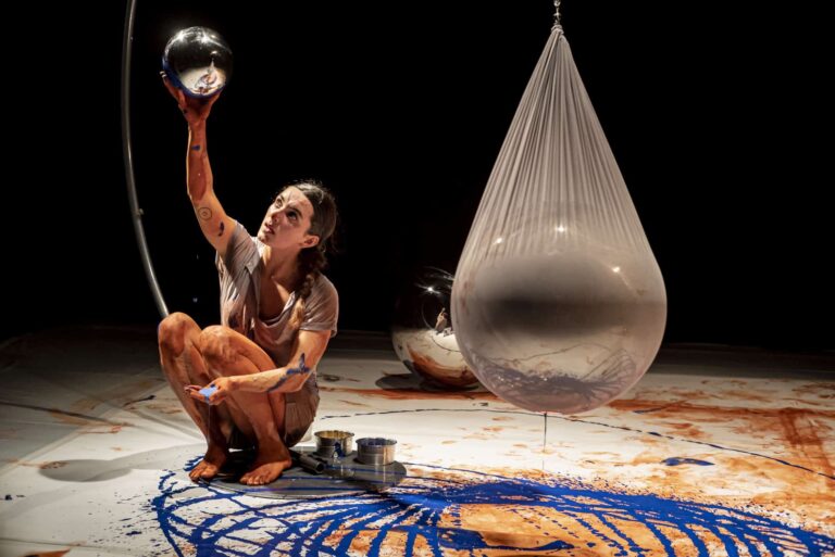 Danser zit op geverfd doek, naast haar hangt een grote plastic zak gevuld met blauwe verf