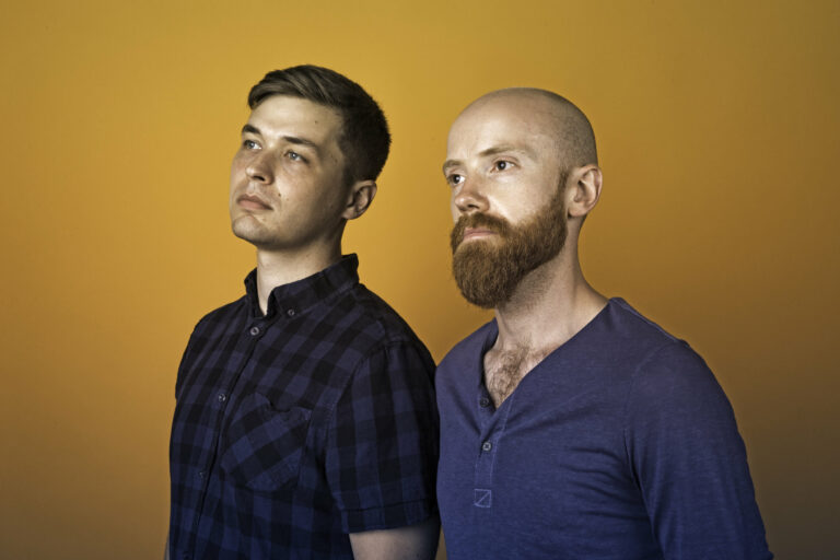 Twee mannen staan zij aan zij voor een achtergrond in een gele tint.