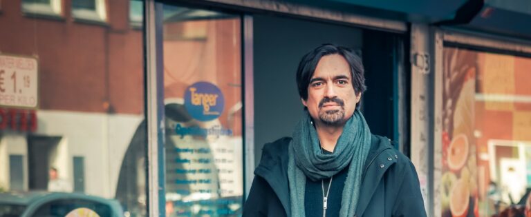 Een man met lang haar en een sjaal staat voor een winkelramen met een logo.