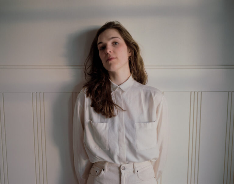 Een jonge vrouw in een witte blouse staat met een neutrale uitdrukking tegen een lichte muur.