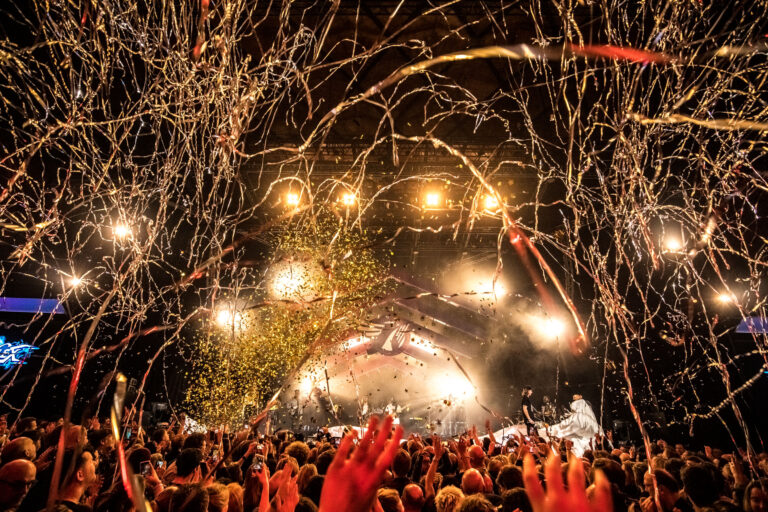 Een levendige concertomgeving met een groot publiek, feestelijke confetti en fel licht van het podium.
