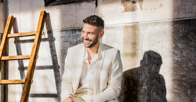 Een portretfoto van Rolf Sanchez die een volledig wit pak draagt en in de zon zit voor een oude, betonnen muur.