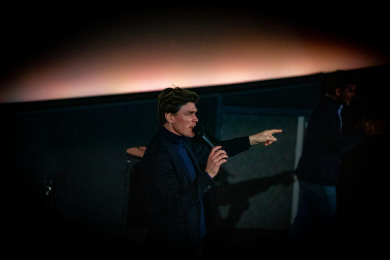 Ingezoomde foto van een man die in een microfoon aan het praten is en de ruimte in wijst.