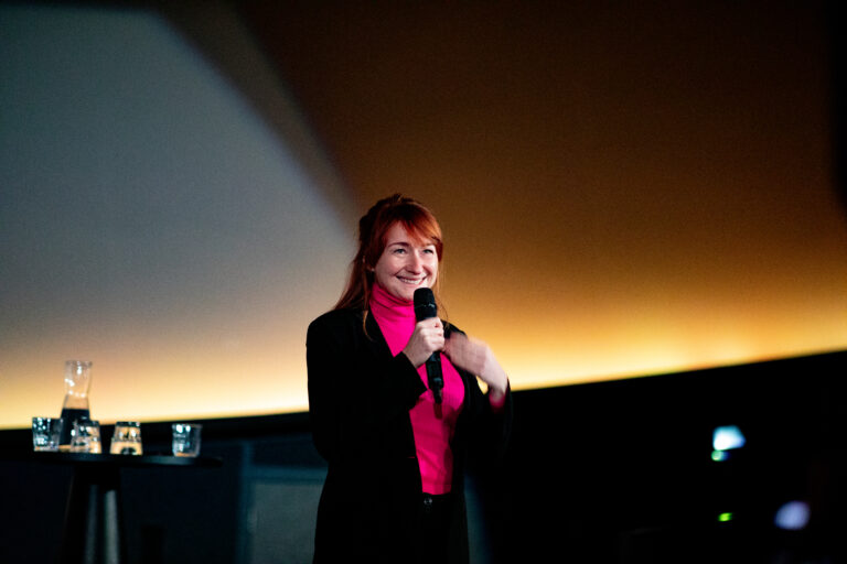 Lachende roodharige vrouw met een fel roze shirt aan en een zwarte blazer erover. Ze heeft een microfoon in haar hand.