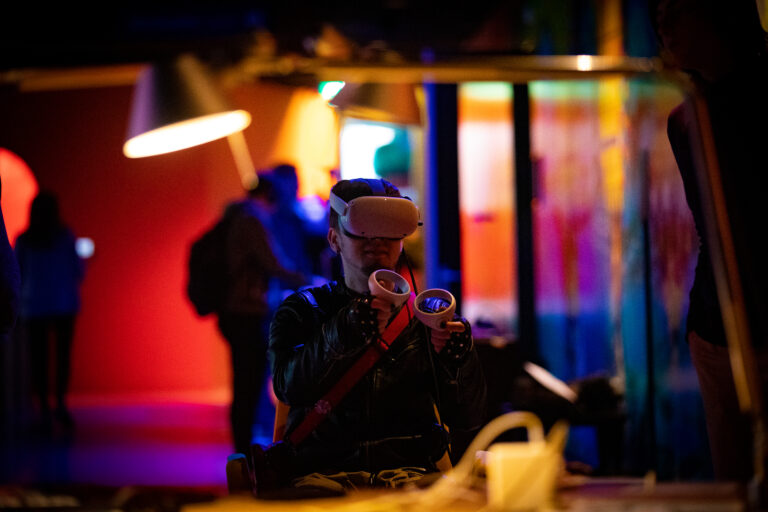 Persoon met een grote witte bril op en twee dingen in de hand. Voor een kleurrijke achtergrond.