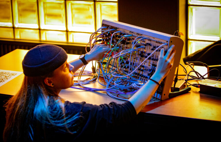 Persoon met een muts op en lang haar die geknield met zijn handen aan een kastje met allemaal stekkertjes zit.