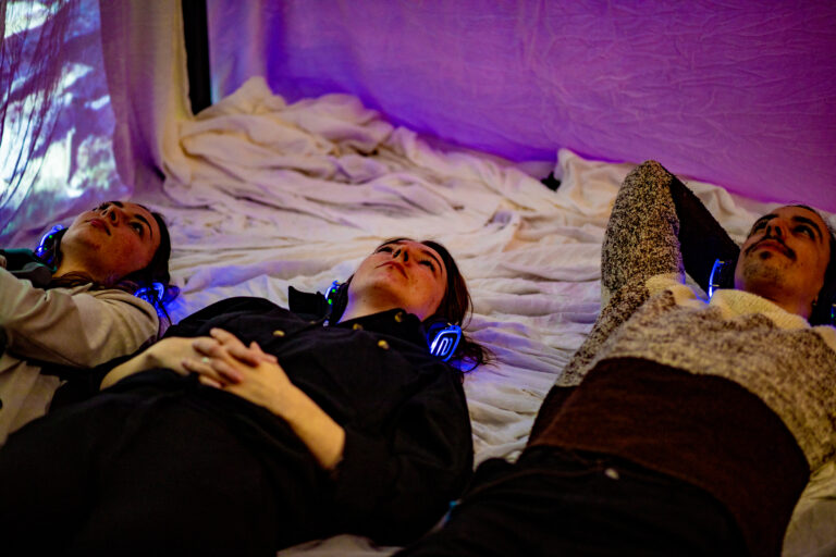 Drie personen die op een bed liggen met hun handen onder hun hoofd, met een koptelefoon op.