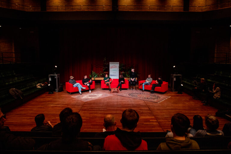 drie rode banken waar per bank twee mensen op zitten, op een podium.