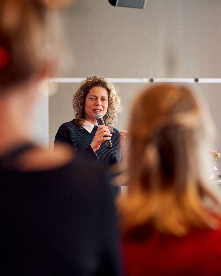 Een spreker met krullend haar staat voor een groep mensen en spreekt in een zaal.