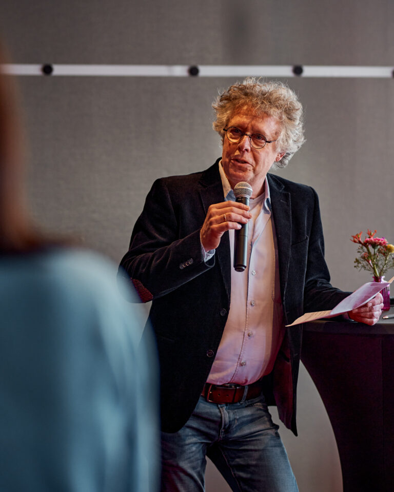 Een man met een witblond haar, gekleed in een zwart jasje, spreekt in een microfoon tijdens een evenement.
