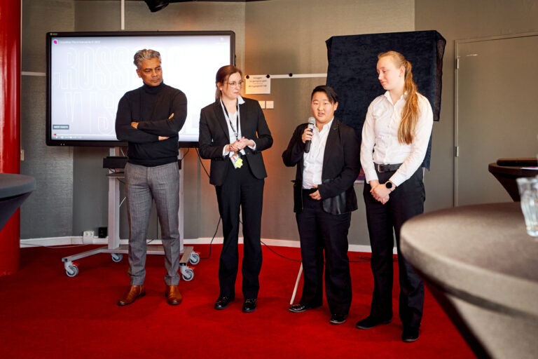 Een groep van vier personen staat op een podium, waarbij één persoon een presentatie geeft en de anderen aandachtig luisteren.