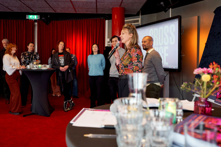 Een groep mensen staat tijdens een presentatie in een zaal met een rode achtergrond, terwijl een spreker het publiek toespreekt.