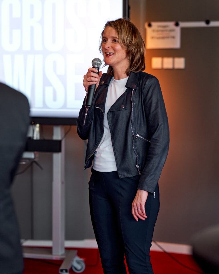 Een vrouw in een leren jas houdt een microfoon vast terwijl ze spreekt op een evenement.