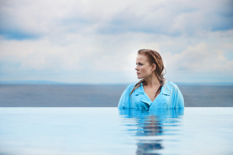 Een foto van Ilse DeLange die naar de zijkant kijkt. Ze zit met een blauw overhemd aan in een buitenzwembad en is helemaal doorweekt.