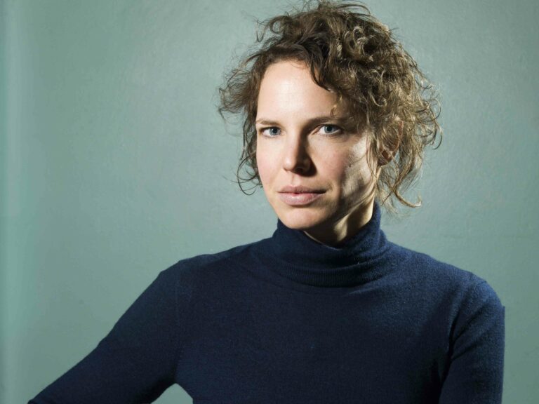 Een vrouw met krullend haar, gekleed in een donkere trui, kijkt serieus in de camera tegen een groene achtergrond.