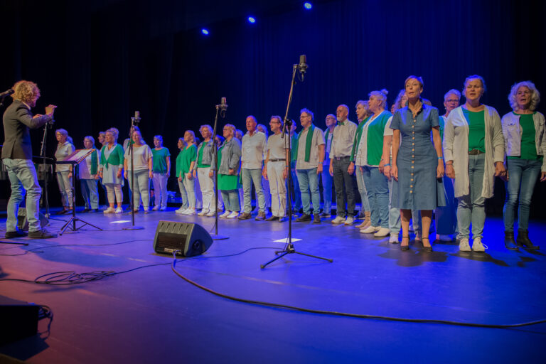 Een koor bestaande uit meerdere zangers staat op een podium met een blauwe achtergrond en microfoons.