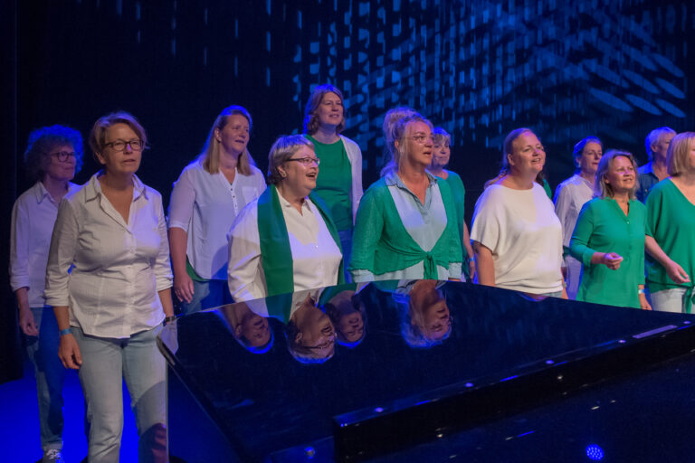 Een koor van vrouwen zingt op een podium met een zwarte piano, omgeven door een blauwe lichtshow.
